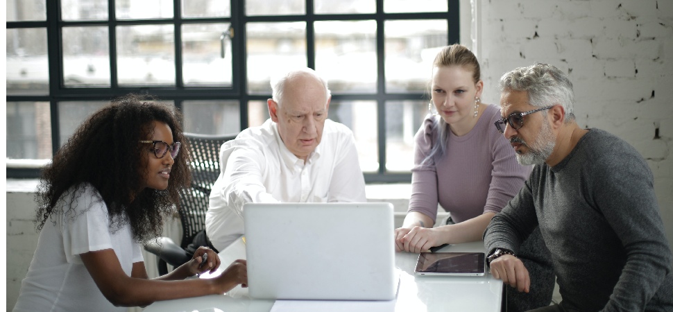 Farklı yaş gruplarından dört profesyonelin birlikte oturup bir dizüstü bilgisayara baktığı, yaşa duyarlı bir iş ortamını gösteren görsel