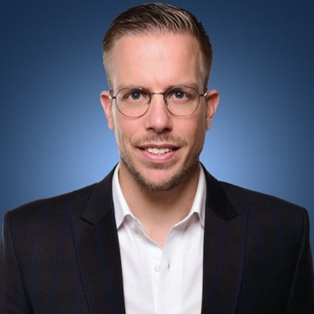 A man wearing glasses and a suit smiles against a blue background.