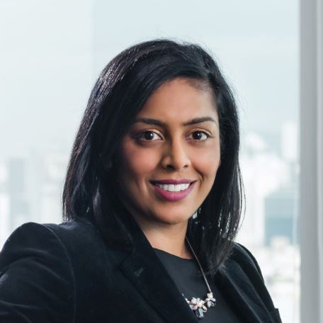 A woman with black hair in business attire smiling in front of a blurred cityscape.