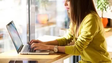 Woman in yellow sweater working on a laptop by a window.