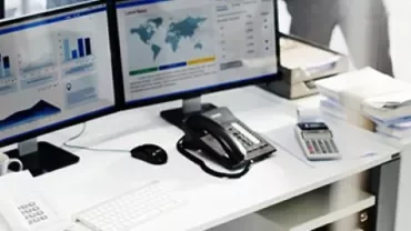 Office desk with two monitors displaying graphs, a telephone, and calculator.