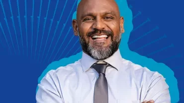 Smiling man in a light shirt and tie against a blue background.