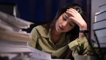 A woman looks tired while reviewing documents at a desk, head in hand.