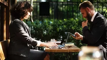 Dışarıda kafede oturan iş kıyafetli iki kişi, laptop ve telefon kullanıyor.
