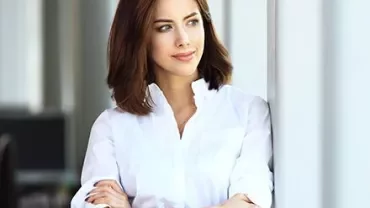 Woman in a white shirt stands thoughtfully by a window.