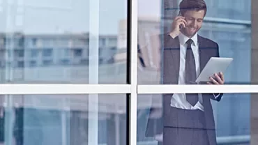 Man in a suit holding a tablet and talking on a phone, standing by a window.