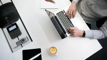 Person typing on a laptop at a desk with a coffee and notepad.