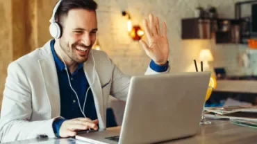 Man smiling and waving at laptop while wearing headphones.