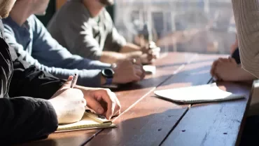People sitting at a table, holding notebooks and pens in a meeting.