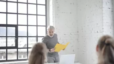 A man with a yellow folder speaking in front of a laptop.