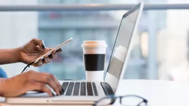 Person holding a phone and working on a laptop with coffee cup nearby.