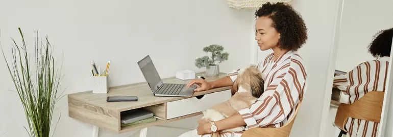 Kadın evde laptopta çalışırken köpeğini kucaklıyor.