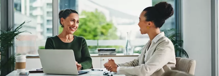 Two women engaged in discussion at a table with a laptop open between them.