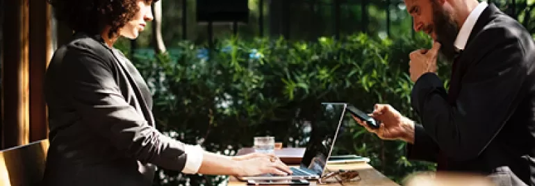 Dışarıda kafede oturan iş kıyafetli iki kişi, laptop ve telefon kullanıyor.