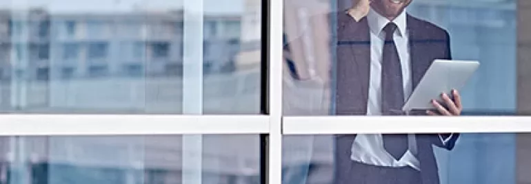 Man in a suit holding a tablet and talking on a phone, standing by a window.