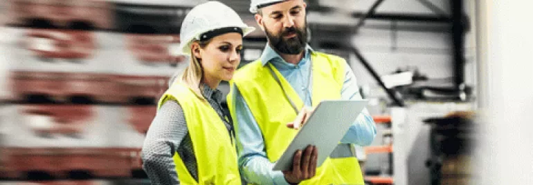 İki inşaat çalışanı, güvenlik ekipmanlarıyla tablet inceliyor.