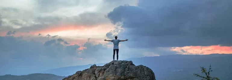 Bir adam, gün batımında bir kayanın üzerinde, kollarını açık bir şekilde duruyor.