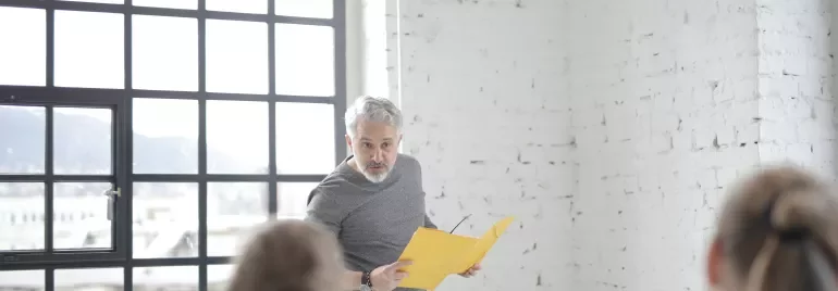 A man with a yellow folder speaking in front of a laptop.