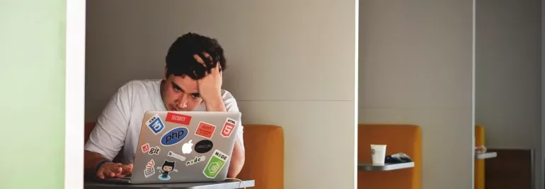 Bilgisayar başında düşünen adam, üzerinde yazılım çıkartmaları olan laptop.