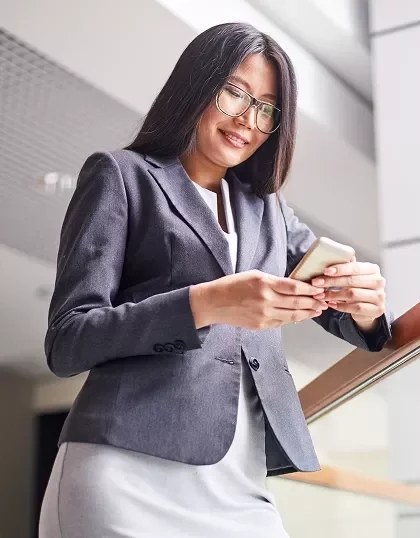 A woman with glasses on a gray suit holding her phone while smiling