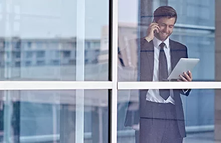 Man in a suit holding a tablet and talking on a phone, standing by a window.